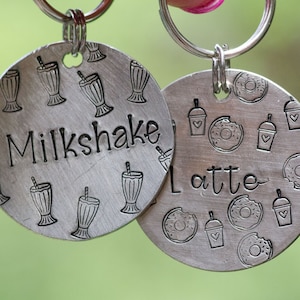 Puede incluir: Dos llaveros redondos de color plateado. Uno tiene la palabra "Milkshake" e ilustraciones de batidos. El otro tiene la palabra "Latte" e ilustraciones de donuts y tazas de café. Ambos llaveros están unidos a anillas plateadas.