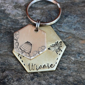 May include: A layered, hexagonal keychain with a silver ring. The top layer is engraved with a beehive and floral design. The bottom layer features a bee and the name "Winnie".