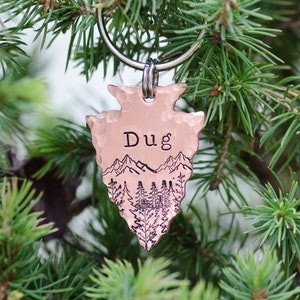 May include: A copper arrowhead-shaped dog tag with the name "Dug" engraved on it. The tag features a mountain and forest scene with a silver ring.