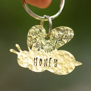 May include: Gold bee-shaped dog tag with a floral design and the word "Honey" engraved on it. The tag is attached to a silver key ring.