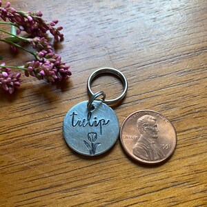 Puede incluir: Una chapa de perro de metal plateado con la palabra "tulip" grabada, un pequeño diseño de tulipán y un anillo plateado. Un centavo de cobre está al lado de la chapa.