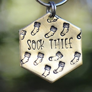 May include: A brass-coloured, hexagonal dog tag with the words "SOCK THIEF" etched in the centre. The tag is adorned with small sock illustrations. A silver ring is attached at the top for easy attachment to a collar.