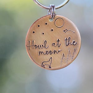 Puede incluir: Una chapa redonda de color bronce con la frase "Howl at the moon". La chapa presenta una silueta de lobo aullando, estrellas, una luna creciente y árboles. Un anillo plateado está unido en la parte superior.