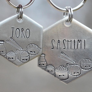K&ouml;nnte beinhalten: Zwei silberfarbene, sechseckige Haustieranh&auml;nger mit Schl&uuml;sselringen. Auf einem Anh&auml;nger steht "TORO", auf dem anderen "SASHIMI". Beide Anh&auml;nger zeigen eingravierte Sushi-Illustrationen, darunter Sojasaucenflaschen, Sushi-Rollen und Essst&auml;bchen.