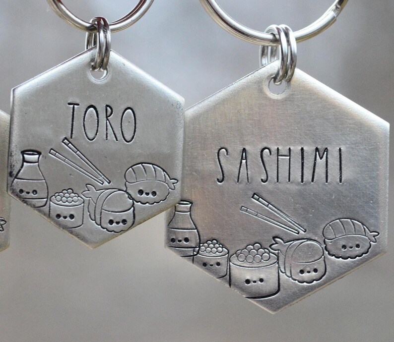 K&ouml;nnte beinhalten: Zwei silberfarbene, sechseckige Haustieranh&auml;nger mit Schl&uuml;sselringen. Auf einem Anh&auml;nger steht "TORO", auf dem anderen "SASHIMI". Beide Anh&auml;nger zeigen eingravierte Sushi-Illustrationen, darunter Sojasaucenflaschen, Sushi-Rollen und Essst&auml;bchen.