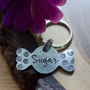 May include: Silver candy-shaped dog tag with the word "Sugar" engraved on it. The tag is attached to a silver key ring.