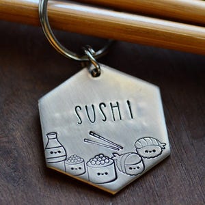 K&ouml;nnte beinhalten: Ein sechseckiger Metall-Schl&uuml;sselanh&auml;nger mit dem eingravierten Wort "SUSHI". Der Schl&uuml;sselanh&auml;nger zeigt eingravierte Illustrationen von Sushi-Rollen, Sojasauce und Essst&auml;bchen. Der Schl&uuml;sselanh&auml;nger ist an einem Metallring befestigt.