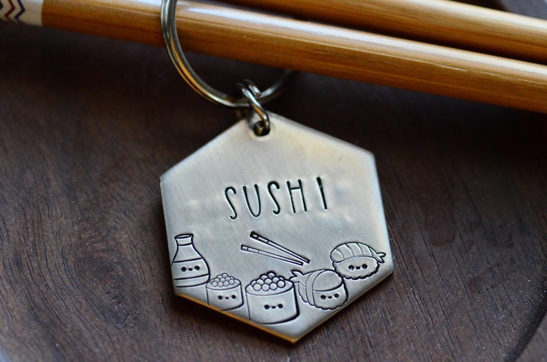 K&ouml;nnte beinhalten: Ein sechseckiger Metall-Schl&uuml;sselanh&auml;nger mit dem eingravierten Wort "SUSHI". Der Schl&uuml;sselanh&auml;nger zeigt eingravierte Illustrationen von Sushi-Rollen, Sojasauce und Essst&auml;bchen. Der Schl&uuml;sselanh&auml;nger ist an einem Metallring befestigt.