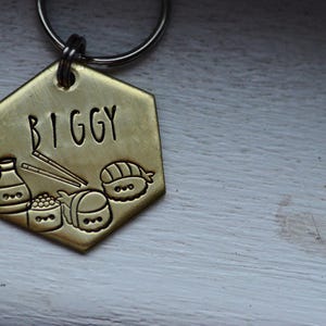 K&ouml;nnte beinhalten: Ein goldfarbener, sechseckiger Hundeanh&auml;nger mit dem Namen "BIGGY" in schwarzer Schrift. Der Anh&auml;nger zeigt ge&auml;tzte Illustrationen von Sushi, Sojasauce und Essst&auml;bchen. Oben ist ein silberner Ring zur Befestigung am Halsband angebracht.