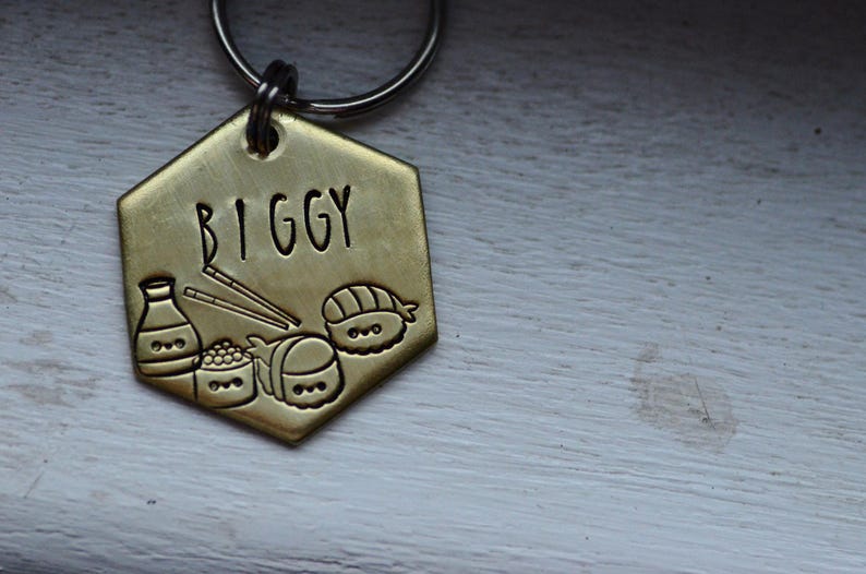 K&ouml;nnte beinhalten: Ein goldfarbener, sechseckiger Hundeanh&auml;nger mit dem Namen "BIGGY" in schwarzer Schrift. Der Anh&auml;nger zeigt ge&auml;tzte Illustrationen von Sushi, Sojasauce und Essst&auml;bchen. Oben ist ein silberner Ring zur Befestigung am Halsband angebracht.