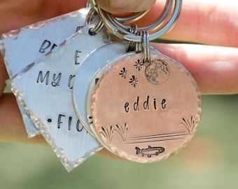 May include: A collection of personalised keyrings with stamped text and designs. A copper-coloured round keyring features the name "eddie", a moon, stars, and a fish. Two silver-coloured keyrings have text. All are attached to a silver keyring.