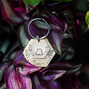 Puede incluir: Un llavero hexagonal de color lat&oacute;n con un dise&ntilde;o de abejas. El llavero presenta una colmena, flores y abejas, con la palabra "Bee" inscrita debajo. Est&aacute; unido a un llavero de color plateado.