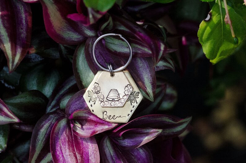 Puede incluir: Un llavero hexagonal de color lat&oacute;n con un dise&ntilde;o de abejas. El llavero presenta una colmena, flores y abejas, con la palabra "Bee" inscrita debajo. Est&aacute; unido a un llavero de color plateado.