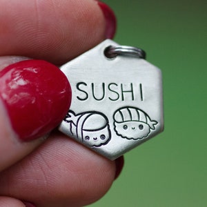 K&ouml;nnte beinhalten: Ein silberfarbener, sechseckiger Anh&auml;nger mit dem eingravierten Wort "SUSHI". Unter dem Wort befinden sich zwei Cartoon-Sushi-Illustrationen. Der Anh&auml;nger hat oben einen kleinen Ring zur Befestigung.