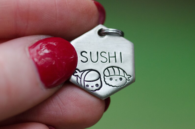K&ouml;nnte beinhalten: Ein silberfarbener, sechseckiger Anh&auml;nger mit dem eingravierten Wort "SUSHI". Unter dem Wort befinden sich zwei Cartoon-Sushi-Illustrationen. Der Anh&auml;nger hat oben einen kleinen Ring zur Befestigung.