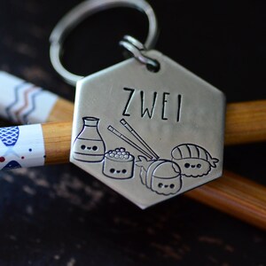 K&ouml;nnte beinhalten: Ein silberfarbener Schl&uuml;sselanh&auml;nger mit dem eingravierten Wort "ZWEI" sowie Illustrationen von Sushi, Sojasauce und Essst&auml;bchen. Der Schl&uuml;sselanh&auml;nger liegt auf einem Paar Essst&auml;bchen und einem dekorativen Essst&auml;bchen.