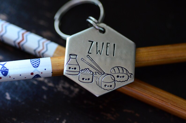 K&ouml;nnte beinhalten: Ein silberfarbener Schl&uuml;sselanh&auml;nger mit dem eingravierten Wort "ZWEI" sowie Illustrationen von Sushi, Sojasauce und Essst&auml;bchen. Der Schl&uuml;sselanh&auml;nger liegt auf einem Paar Essst&auml;bchen und einem dekorativen Essst&auml;bchen.
