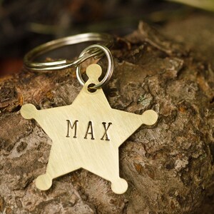 May include: A gold-colored star-shaped dog tag with the name "MAX" engraved on it. The tag is attached to a silver key ring.