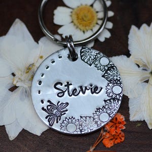 Può includere: Un portachiavi rotondo in metallo color argento con il nome "Stevie" in corsivo nero. Il tag presenta un design di api e fiori. Lo sfondo include fiori bianchi e una margherita.