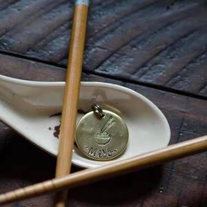 K&ouml;nnte beinhalten: Ein Messinganh&auml;nger mit einem Nudelsch&uuml;ssel- und Essst&auml;bchen-Design, der auf einem wei&szlig;en Keramik-L&ouml;ffel liegt. Der Anh&auml;nger tr&auml;gt die Gravur "Miso". Zwei Bambus-Essst&auml;bchen sind um den L&ouml;ffel auf einer dunklen Holzoberfl&auml;che angeordnet.