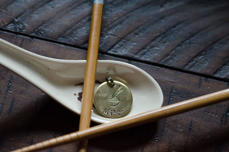 K&ouml;nnte beinhalten: Ein Messinganh&auml;nger mit einem Nudelsch&uuml;ssel- und Essst&auml;bchen-Design, der auf einem wei&szlig;en Keramik-L&ouml;ffel liegt. Der Anh&auml;nger tr&auml;gt die Gravur "Miso". Zwei Bambus-Essst&auml;bchen sind um den L&ouml;ffel auf einer dunklen Holzoberfl&auml;che angeordnet.