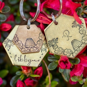 Puede incluir: Dos llaveros hexagonales de lat&oacute;n con dise&ntilde;os de abejas y flores. Un llavero tiene grabado el nombre "libby", y el otro la letra "R". Los llaveros cuelgan de un anillo plateado, con un fondo de flores rojas y hojas verdes.