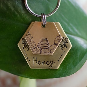 Puede incluir: Una etiqueta de llavero de color lat&oacute;n con un dise&ntilde;o de panal. La etiqueta presenta ilustraciones de colmena y flores, junto con dos abejas. La palabra "Honey" est&aacute; inscrita debajo. La etiqueta est&aacute; unida a un llavero plateado.