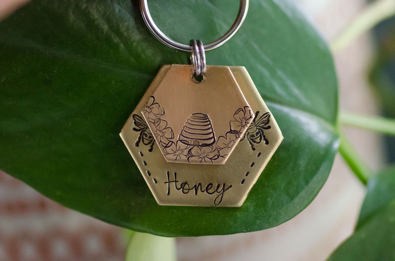Puede incluir: Una etiqueta de llavero de color lat&oacute;n con un dise&ntilde;o de panal. La etiqueta presenta ilustraciones de colmena y flores, junto con dos abejas. La palabra "Honey" est&aacute; inscrita debajo. La etiqueta est&aacute; unida a un llavero plateado.