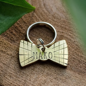 May include: A gold-colored bowtie-shaped dog tag with the name "MAKO" engraved on it. The tag is attached to a silver key ring.