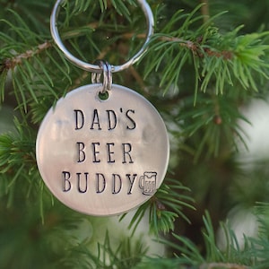 May include: A circular, silver-toned keychain tag with the words "DAD'S BEER BUDDY" stamped on it. A small beer mug graphic is below the text. The tag hangs from a silver keyring, set against a backdrop of green pine needles.