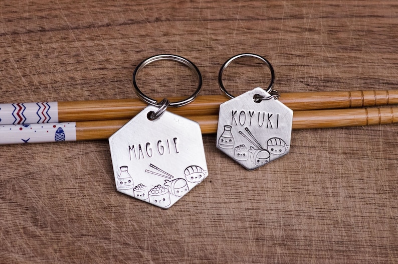 K&ouml;nnte beinhalten: Zwei silberne sechseckige Schl&uuml;sselanh&auml;nger mit den eingravierten Namen "MAGGIE" und "KOYUKI". Jeder Schl&uuml;sselanh&auml;nger hat ein Sushi-Motiv mit Sojasaucenflaschen, Sushi-Rollen und Essst&auml;bchen. Die Schl&uuml;sselanh&auml;nger liegen auf einer Holzoberfl&auml;che mit Essst&auml;bchen.