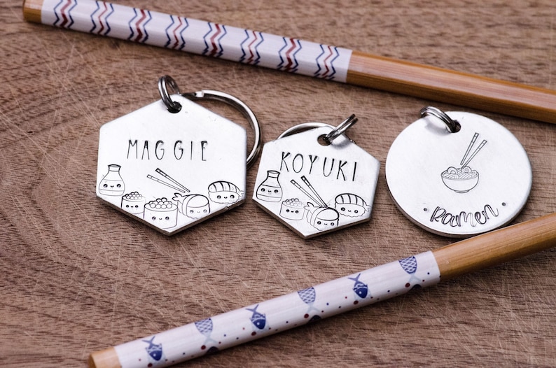 K&ouml;nnte beinhalten: Silberne Schl&uuml;sselanh&auml;nger mit Sushi- und Ramen-Designs. Zwei Schl&uuml;sselanh&auml;nger sind sechseckig und mit Namen und Sushi-Illustrationen graviert. Ein runder Schl&uuml;sselanh&auml;nger zeigt eine Ramen-Sch&uuml;ssel. Essst&auml;bchen mit dekorativen Papierb&auml;ndern im Hintergrund.