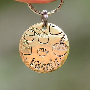 K&ouml;nnte beinhalten: Ein runder, goldfarbener Hundeanh&auml;nger mit geh&auml;mmertem Muster. Der Anh&auml;nger zeigt ge&auml;tzte Illustrationen von Sushi, Ramen und Essst&auml;bchen. Das Wort "Kimchi" ist in Schreibschrift unter den Essensillustrationen geschrieben.