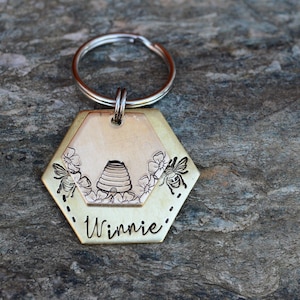 Puede incluir: Un llavero hexagonal de color lat&oacute;n con un anillo plateado. El llavero presenta un dise&ntilde;o con tem&aacute;tica de abejas con una colmena, flores y abejas. El nombre "Winnie" est&aacute; grabado en la parte inferior. El fondo es una superficie de piedra gris texturizada.