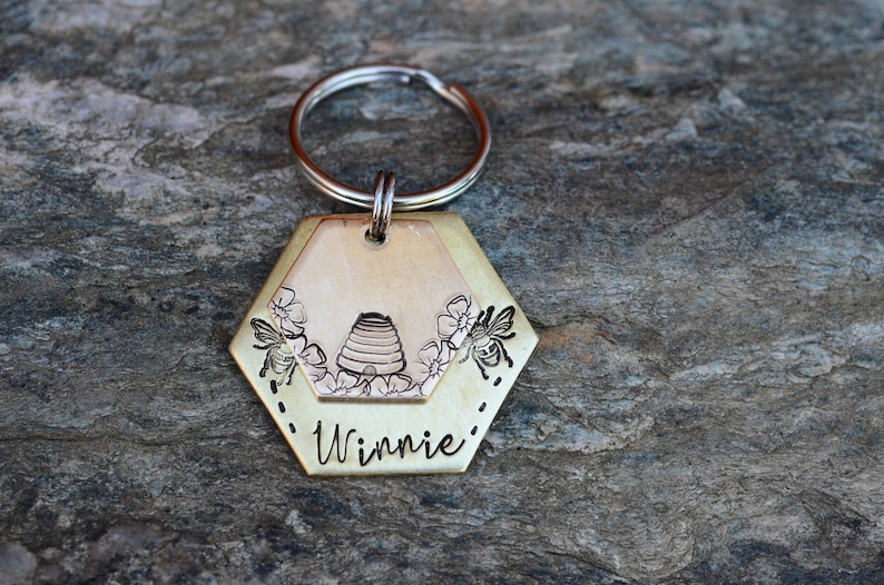Puede incluir: Un llavero hexagonal de color lat&oacute;n con un anillo plateado. El llavero presenta un dise&ntilde;o con tem&aacute;tica de abejas con una colmena, flores y abejas. El nombre "Winnie" est&aacute; grabado en la parte inferior. El fondo es una superficie de piedra gris texturizada.