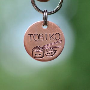 K&ouml;nnte beinhalten: Eine runde, kupferfarbene Haustier-Marke mit dem Wort "TOBIKO" oben eingraviert. Darunter befinden sich Illustrationen von Sushi und Essst&auml;bchen. Die Marke ist an einem silberfarbenen Ring befestigt.