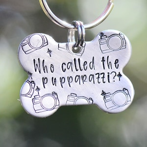 Puede incluir: Una placa de identificación para perros plateada con forma de hueso, con la frase "Who called the pupparazzi?" en letras negras. La placa está adornada con ilustraciones de cámaras y estrellas, y está sujeta a un anillo plateado.