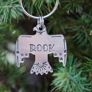 May include: A silver-colored metal pet tag in the shape of a stylized bird with the word "ROOK" engraved on it. The tag is attached to a silver keyring and is set against a backdrop of green foliage.