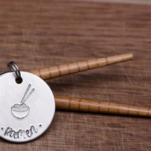 K&ouml;nnte beinhalten: Ein runder, silberfarbener Anh&auml;nger mit einem gestempelten Ramen-Sch&uuml;ssel- und Essst&auml;bchen-Design. Das Wort "Ramen" ist darunter gestempelt. Zwei Bambus-Essst&auml;bchen liegen hinter dem Anh&auml;nger auf einer Holzoberfl&auml;che.