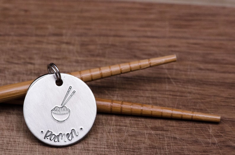 K&ouml;nnte beinhalten: Ein runder, silberfarbener Anh&auml;nger mit einem gestempelten Ramen-Sch&uuml;ssel- und Essst&auml;bchen-Design. Das Wort "Ramen" ist darunter gestempelt. Zwei Bambus-Essst&auml;bchen liegen hinter dem Anh&auml;nger auf einer Holzoberfl&auml;che.