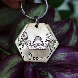 Puede incluir: Un llavero de color lat&oacute;n con una etiqueta hexagonal con un dise&ntilde;o de abeja. La etiqueta tiene una abeja, flores y una ilustraci&oacute;n de colmena. La palabra "Bee" est&aacute; escrita debajo de la colmena. El llavero est&aacute; sujeto a un anillo plateado.