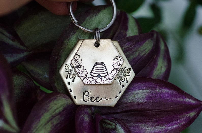 Puede incluir: Un llavero de color lat&oacute;n con una etiqueta hexagonal con un dise&ntilde;o de abeja. La etiqueta tiene una abeja, flores y una ilustraci&oacute;n de colmena. La palabra "Bee" est&aacute; escrita debajo de la colmena. El llavero est&aacute; sujeto a un anillo plateado.