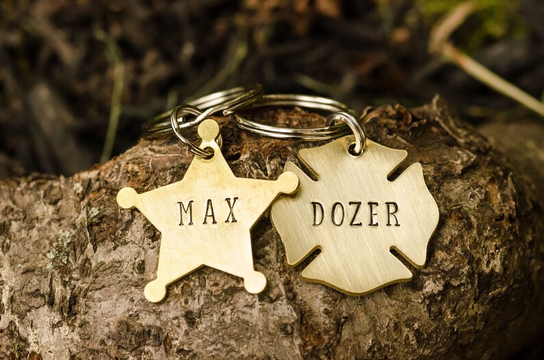 May include: Two brass dog tags on a key ring. One tag is a star shape with the name "MAX" engraved on it. The other tag is a fire hydrant shape with the name "DOZER" engraved on it.