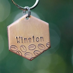 May include: A copper-colored, hexagonal pet tag with the name "Winston" stamped on it. The bottom edge features an etched design of coffee beans. The tag is attached to a silver keyring, perfect for a pet.