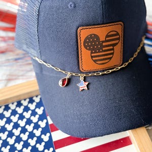 Puede incluir: Una gorra de camionero azul marino con un parche de cuero tostado rectangular que presenta una silueta de Mickey Mouse grabada con láser, rellena de estrellas y rayas. Una cadena dorada rodea la parte frontal con un dije de gota rojo y una estrella.