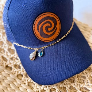 May include: A navy blue baseball cap featuring a circular brown leather patch with a dark brown spiral design. A gold-toned chain is draped across the front, adorned with a small white seashell charm and a teardrop-shaped blue crystal pendant.