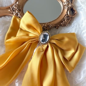 May include: A golden yellow satin bow with a cameo brooch and a small dangling charm. The bow is set against a gold-framed hand mirror with a floral design. The background is a white lace fabric.