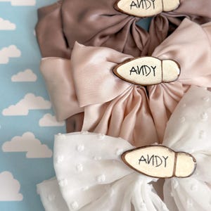 Puede incluir: Tres lazos para el pelo de tela en tonos marrón, rosa y blanco. Cada lazo presenta una pieza de acento de madera con el nombre "ANDY" en letras negras. Los lazos están dispuestos sobre un fondo azul claro con formas de nubes blancas.