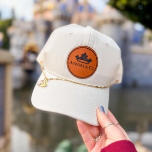 May include: Cream-colored baseball cap with a brown leather patch featuring a crown logo and the text "AURORA & CO." A gold chain with a charm is attached to the brim. The cap is held up in front of a blurred background.