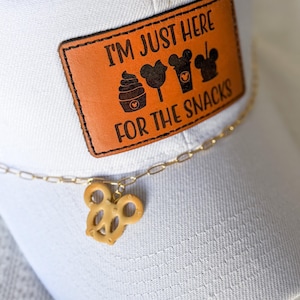 May include: White baseball cap with a brown leather patch that reads "I'M JUST HERE FOR THE SNACKS" with food icons. A gold chain with a pretzel charm hangs from the cap.